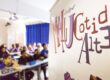 Sala de aula com estudantes ao fundo e parede com texto "Oficina Maluca" em destaque
